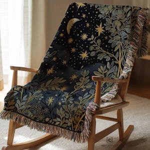 May include: A dark blue throw blanket draped over a wooden rocking chair. The blanket features a celestial design with a crescent moon, stars, and botanical elements in gold and green. The blanket has a fringed edge.