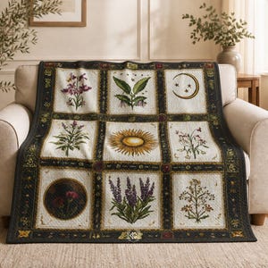 May include: A decorative throw blanket with a patchwork design featuring botanical illustrations and celestial motifs. The quilt displays images of flowers, a sun, a moon, and stars, framed within a dark border. The blanket is draped over a beige armchair.