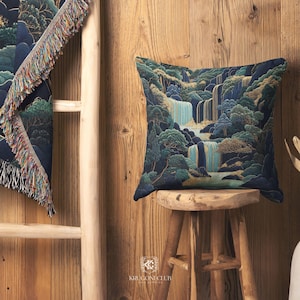 May include: A square decorative pillow with a waterfall landscape design in shades of blue, green, and gold. The pillow is placed on a small wooden stool. A matching throw blanket with fringe is partially visible.