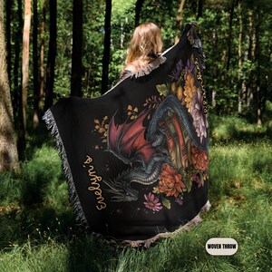 May include: A black woven throw blanket featuring a dragon with red wings perched on a stack of books, surrounded by colorful floral illustrations. The text "Evelyn's" is printed in a script font on the left side of the blanket.