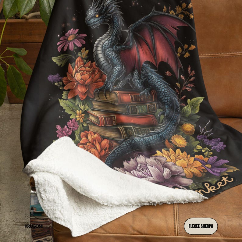 May include: A black throw blanket with a white sherpa lining featuring a printed illustration of a dark dragon perched on a stack of books surrounded by colorful flowers in shades of orange, yellow, and purple. The text FLECEE SHERPA is visible.