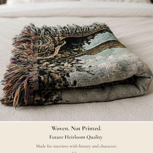 Pode incluir: Uma manta tecida dobrada com uma cena de paisagem detalhada em tons de verde escuro, preto e azul claro. A manta tem uma borda com franjas de fios multicoloridos. O texto na imagem diz: Woven. Not Printed. Future Heirloom Quality. Made for interiors with history and character.