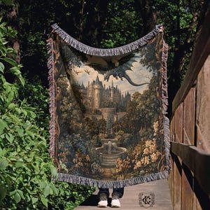 May include: A tapestry throw blanket featuring a fantasy castle scene with a dragon in flight. The blanket has a detailed illustration with a castle, trees, and a fountain. The edges are finished with fringe.