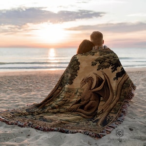 May include: A beige tapestry blanket featuring a detailed illustration of a dragon with outstretched wings, set against a landscape of trees and a forest. The blanket has a fringed edge and is draped over two people on a beach at sunset.