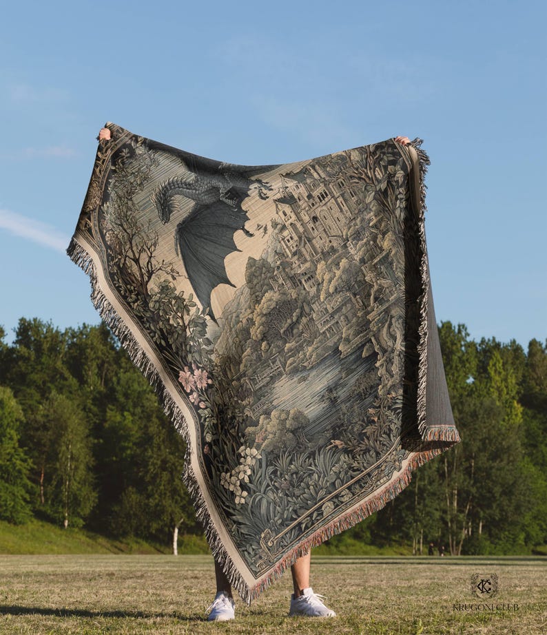 May include: A woven throw blanket featuring a detailed illustration of a large dragon flying over a castle, a lake, and lush foliage. The tapestry-style blanket has a fringed edge and displays a muted color palette of dark greens, browns, and creams.