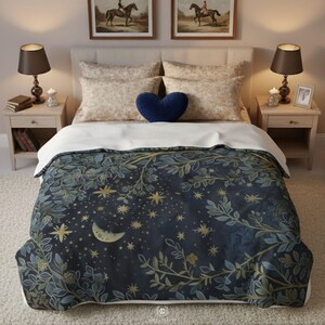 Celestial moon and stars fleece minky blanket displayed on elegant bedroom bed with white linens and navy blue heart pillow, featuring botanical sage green foliage border surrounding crescent moon and golden stars on dark navy background, creating cozy witchy boho bedroom decor perfect for mystical home aesthetics