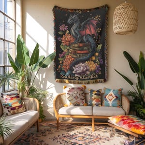 May include: A woven throw blanket featuring a dark background with a dragon perched on a stack of books surrounded by colorful flowers. The text Reading blanket is printed at the bottom. The blanket has fringed edges and a multi-colored border.