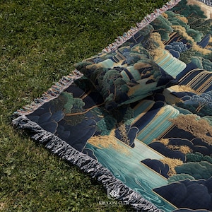 May include: A woven blanket and pillow set with a landscape design featuring trees, water, and mountains in shades of blue, green, and gold. The blanket has a fringed edge and the pillow is square. The blanket and pillow are on a green grassy surface.