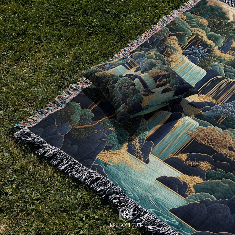 May include: A woven blanket and pillow set with a landscape design featuring trees, water, and mountains in shades of blue, green, and gold. The blanket has a fringed edge and the pillow is square. The blanket and pillow are on a green grassy surface.