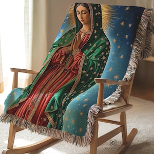 May include: A woven throw blanket featuring a depiction of Our Lady of Guadalupe with a green mantle, red robe, and gold star accents against a blue starry background, finished with decorative fringe edges and draped over a wooden rocking chair.