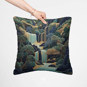 May include: Square decorative pillow featuring a detailed landscape design with waterfalls, trees, and rocks. The color palette includes shades of blue, green, and gold. The pillow is held by a hand.