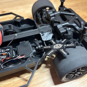 May include: A black remote-controlled car chassis featuring a Spektrum electronic speed controller with red and black wiring, a black plastic suspension arm, and a black star-shaped ride height gauge tool labeled with 1mm, 1.5mm, 2mm, 2.5mm, and 3mm.