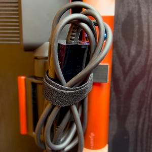 May include: A black electrical cord is wrapped around a black and red hair styling tool. The cord is secured with a black velcro strap. The hair styling tool is mounted on a wall.