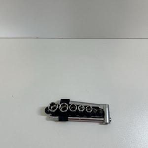 May include: A small, silver and black socket wrench set. The set includes multiple sockets of varying sizes, held together by a black strap. The wrench has a pivoting head for angled access.