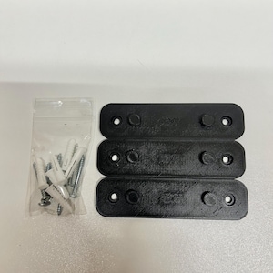 Może przedstawiać: Trzy czarne plastikowe uchwyty ścienne z otworami na śruby i dwa białe plastikowe kołki w przezroczystym plastikowym woreczku.