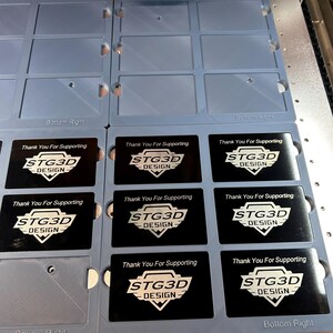 May include: A close-up of a laser cutter engraving business cards with the text "Thank You For Supporting STG3D DESIGN" on each card. The cards are arranged in a grid pattern on a gray surface.
