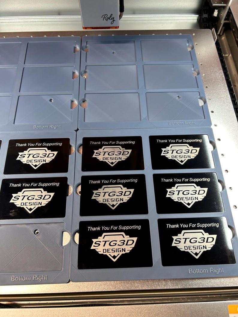 May include: A close-up of a laser cutter engraving business cards with the text "Thank You For Supporting STG3D DESIGN" on each card. The cards are arranged in a grid pattern on a gray surface.