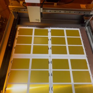 May include: A close-up of a laser cutting machine with a sheet of gold-colored material being cut into 12 rectangular pieces. The machine is labeled "Rolz".
