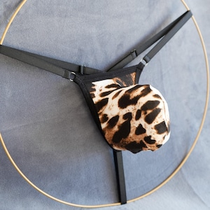 Puede incluir: Un tanga negro y marrón con estampado de leopardo y tirantes ajustables. El tanga se muestra en un anillo dorado.