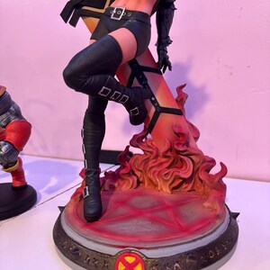May include: A detailed figurine of a female character in a dynamic pose, clad in a black outfit with knee-high boots. The figure stands on a base with a fiery, orange and red design, and a circular emblem.