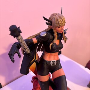 May include: A detailed figurine of a female character in a dynamic pose. She wears a black outfit with a crop top, shorts, and thigh-high boots. The figure has blonde hair and is holding a weapon. The figurine has a detailed design and is on a decorative base.