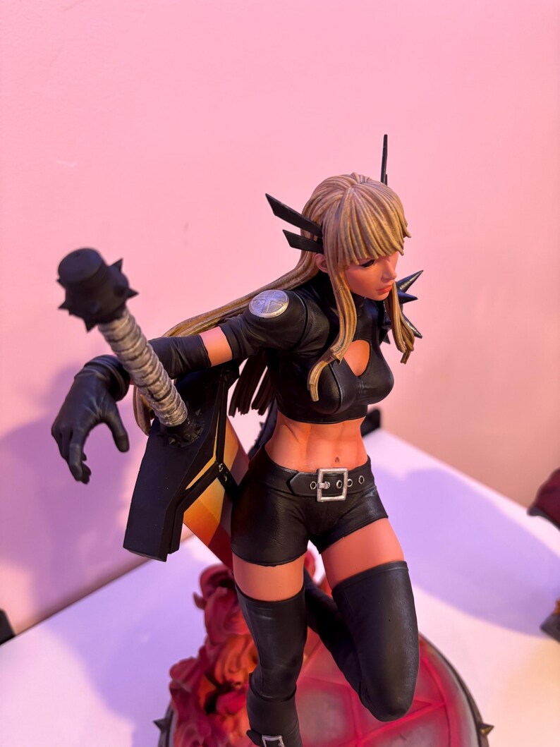 May include: A detailed figurine of a female character in a dynamic pose. She wears a black outfit with a crop top, shorts, and thigh-high boots. The figure has blonde hair and is holding a weapon. The figurine has a detailed design and is on a decorative base.