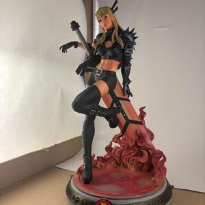 May include: A detailed figurine of a female character in a black outfit, holding a sword. The figure has blonde hair and is posed dynamically on a base with a fiery design. The base features a red and gold color scheme.