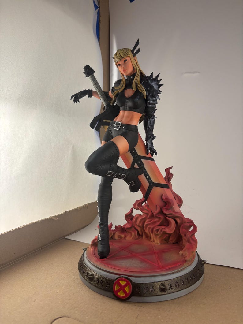 May include: A detailed figurine of a female character in a black outfit, holding a sword. The figure has blonde hair and is posed dynamically on a base with a fiery design. The base features a red and gold color scheme.