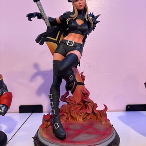 May include: A detailed figurine of a female character in a black outfit, holding a guitar-shaped weapon. The figure stands on a base with flames and a red pentagram. The character has blonde hair and is wearing black boots.