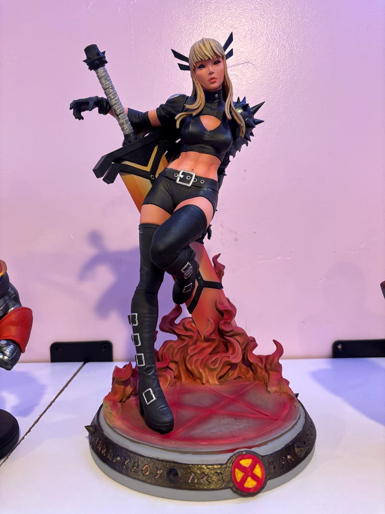 May include: A detailed figurine of a female character in a black outfit, holding a guitar-shaped weapon. The figure stands on a base with flames and a red pentagram. The character has blonde hair and is wearing black boots.