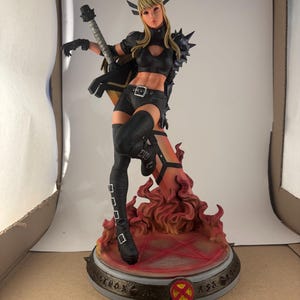 May include: A detailed figurine of a female character in a black outfit, holding a weapon. The figure stands on a base with a fiery design and an X-Men logo. The character has blonde hair and is in a dynamic pose.