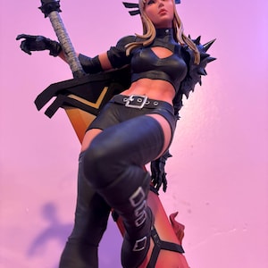May include: A detailed statue of a female character in a black outfit, holding a large sword. The figure has blonde hair and is posed dynamically. The sword has a yellow blade and black handle. The statue is set against a pink background.