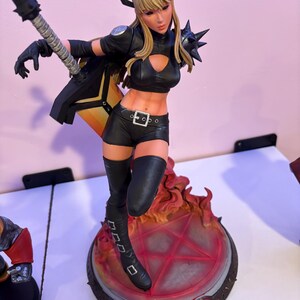 May include: A detailed figurine of a female character in a black outfit, boots, and gloves, with blonde hair and a weapon. The figure stands on a base with a red pentagram design and flames. The figurine has a dynamic pose.