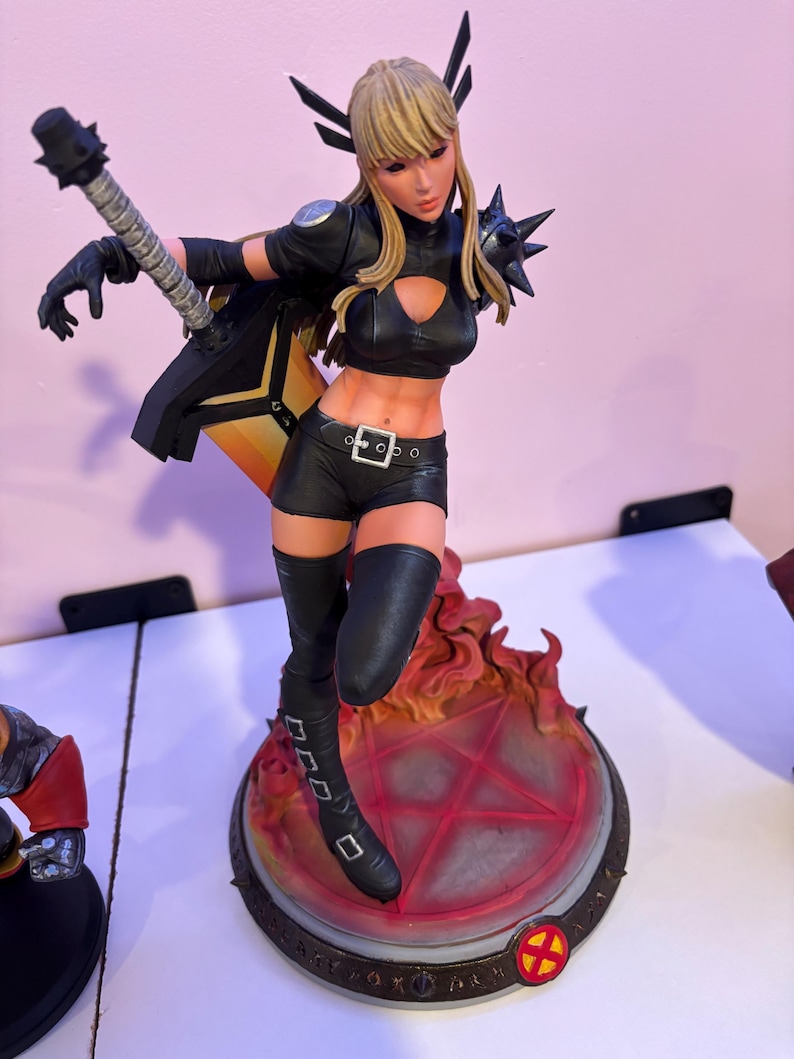 May include: A detailed figurine of a female character in a black outfit, boots, and gloves, with blonde hair and a weapon. The figure stands on a base with a red pentagram design and flames. The figurine has a dynamic pose.