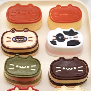 Pode incluir: Uma bandeja com seis sobremesas em forma de gato. As sobremesas têm várias cores, incluindo laranja, branco, preto, castanho e verde. Cada sobremesa apresenta um design de rosto de gato com olhos, nariz e bigodes. As sobremesas são em camadas e parecem pequenos bolos ou pastelaria.