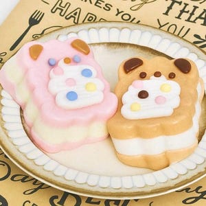 May include: Two decorative, bear-shaped desserts on a plate. One is pink, the other is tan, both with white frosting and colorful sprinkles. The plate has a scalloped edge and sits on a patterned surface. The desserts have bear ears.