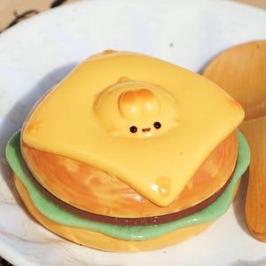 May include: A yellow cheese-like topping covers a green and brown hamburger-shaped object. The topping has a small, yellow, cartoon face with black eyes.
