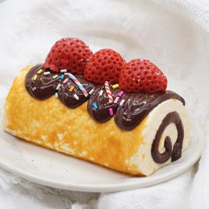 May include: A decorative dessert roll with a cream filling and a chocolate swirl. The roll is topped with chocolate frosting, red strawberries, and colourful sprinkles. It sits on a white plate against a white background.