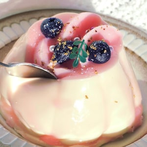 May include: A close-up of a dessert on a plate. The dessert is a light pink and white pudding, topped with blueberries, gold flakes, and a green leaf. A spoon is inserted into the pudding.