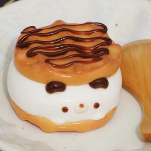 May include: A white and brown waffle ice cream sandwich with chocolate drizzle and a cute cartoon face.