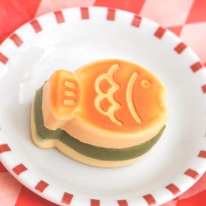 May include: A yellow and orange fish-shaped cake with a green filling on a white plate with a red and white checkered pattern.