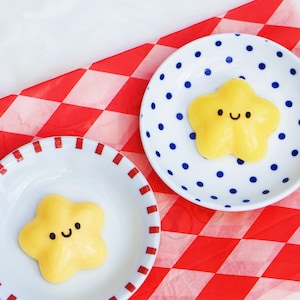 May include: Two yellow star-shaped desserts with smiling faces on white plates with red and blue trim. The plates are on a red and white checked tablecloth.