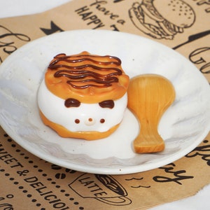 K&ouml;nnte beinhalten: Ein wei&szlig;er Teller mit einem braun-wei&szlig; gestreiften Kuchen mit Schokoladenfrosting und einem Holzl&ouml;ffel. Der Teller liegt auf einem braunen Papieruntergrund mit den aufgedruckten Worten "Have a Happy" und "Latte".