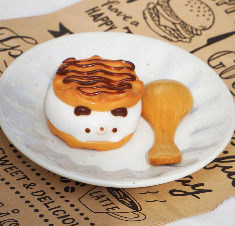 K&ouml;nnte beinhalten: Ein wei&szlig;er Teller mit einem braun-wei&szlig; gestreiften Kuchen mit Schokoladenfrosting und einem Holzl&ouml;ffel. Der Teller liegt auf einem braunen Papieruntergrund mit den aufgedruckten Worten "Have a Happy" und "Latte".