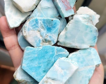 May include: A handful of raw larimar stones, showcasing a range of blue and white hues. The stones have a natural, rough texture, with some displaying reddish-brown inclusions. The color palette varies from light turquoise to deep ocean blue.