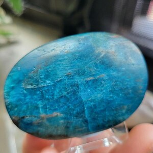 May include: A polished, oval gemstone in shades of turquoise and blue, with natural veining and patterns. The stone is held on a clear acrylic stand, showcasing its smooth, reflective surface. This unique piece is a decorative item.