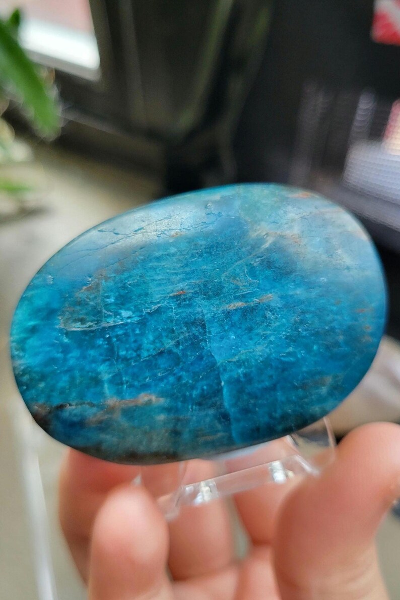 May include: A polished, oval gemstone in shades of turquoise and blue, with natural veining and patterns. The stone is held on a clear acrylic stand, showcasing its smooth, reflective surface. This unique piece is a decorative item.