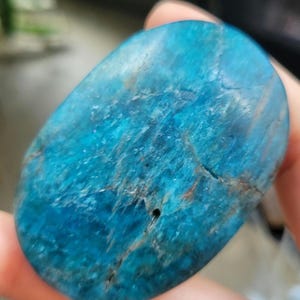 May include: A polished, oval-shaped stone in a vibrant blue colour, with streaks of lighter blue and brown. The stone has a smooth surface with natural imperfections, and a small hole. This gemstone is a natural mineral.