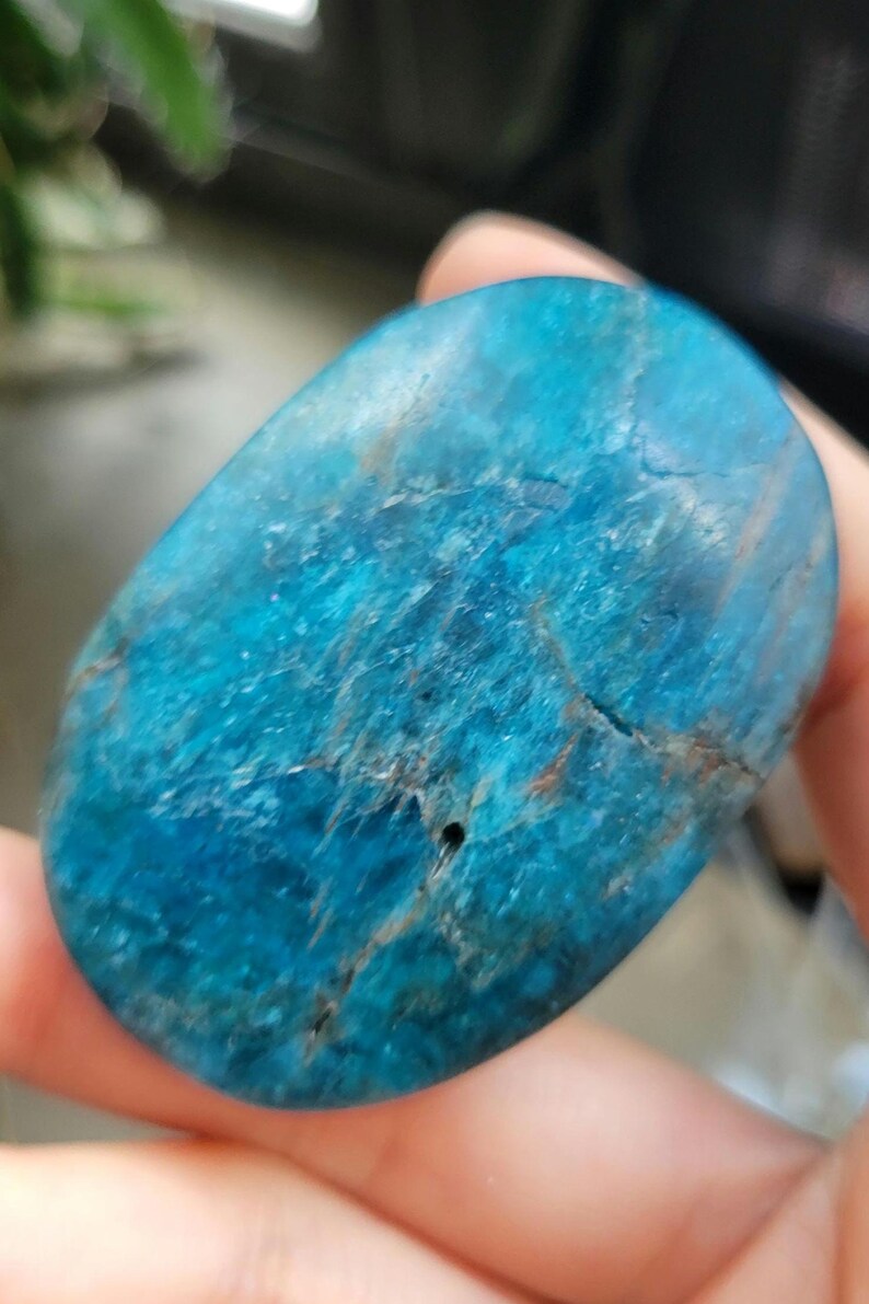 May include: A polished, oval-shaped stone in a vibrant blue colour, with streaks of lighter blue and brown. The stone has a smooth surface with natural imperfections, and a small hole. This gemstone is a natural mineral.