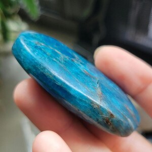 May include: A polished, oval-shaped blue apatite gemstone. The stone has a smooth surface with natural variations in colour and veining. The gemstone is held in a hand, with a blurred background.